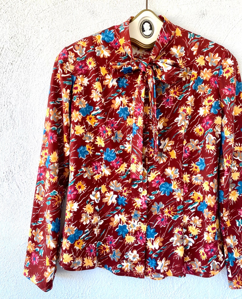 Pode incluir: Uma blusa de manga comprida com um la&ccedil;o no pesco&ccedil;o. A blusa &eacute; feita de um tecido marrom com uma estampa floral em tons de vermelho, azul, amarelo e branco.