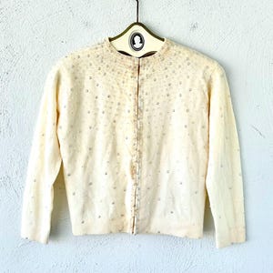 May include: Cream-colored vintage cardigan with long sleeves, adorned with scattered silver-colored beads. The sweater is buttoned up and hangs on a hanger against a white wall. The cardigan has a round neckline.