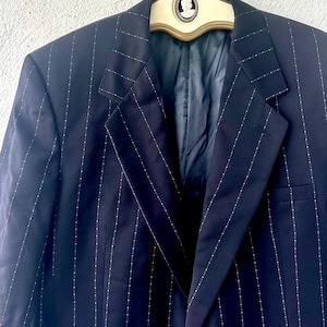 Peut inclure: Un blazer bleu marine fonc&eacute; pr&eacute;sentant un motif &agrave; rayures verticales avec de petits points blancs. Le blazer poss&egrave;de des revers crant&eacute;s et est pr&eacute;sent&eacute; sur un cintre cr&egrave;me orn&eacute; d'un embl&egrave;me cam&eacute;e ovale central avec une silhouette de profil blanche.