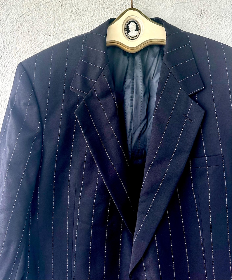 Peut inclure: Un blazer bleu marine fonc&eacute; pr&eacute;sentant un motif &agrave; rayures verticales avec de petits points blancs. Le blazer poss&egrave;de des revers crant&eacute;s et est pr&eacute;sent&eacute; sur un cintre cr&egrave;me orn&eacute; d'un embl&egrave;me cam&eacute;e ovale central avec une silhouette de profil blanche.