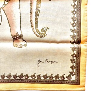 May include: A cream-colored silk scarf featuring a detailed illustration of an elephant with ornate detailing. The scarf has a decorative border and the signature "Jim Thompson" in the corner. The elephant is in shades of brown and gold.