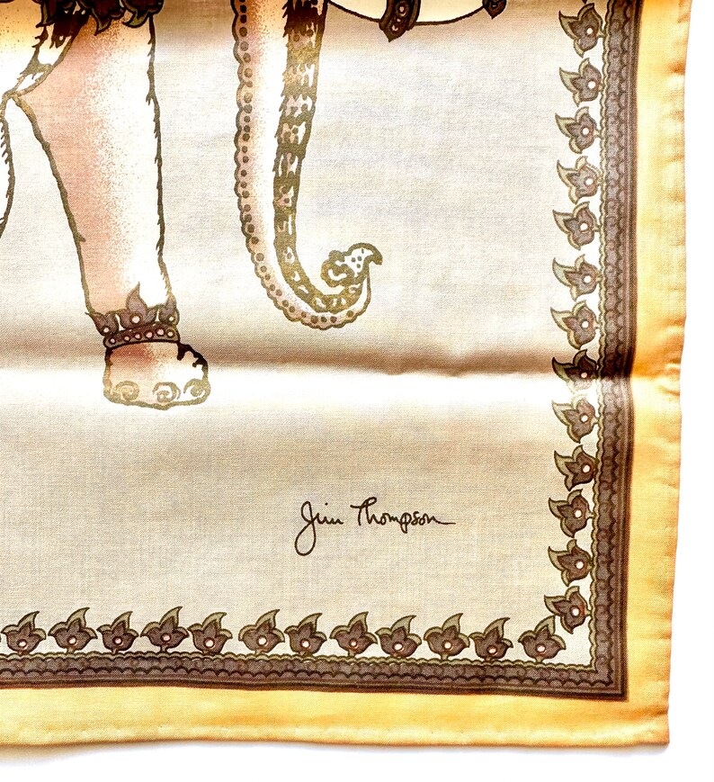 May include: A cream-colored silk scarf featuring a detailed illustration of an elephant with ornate detailing. The scarf has a decorative border and the signature "Jim Thompson" in the corner. The elephant is in shades of brown and gold.