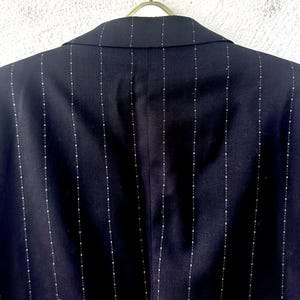 Peut inclure: Un blazer bleu marine fonc&eacute; pr&eacute;sentant un motif &agrave; rayures verticales compos&eacute; de petites lignes pointill&eacute;es claires. Le v&ecirc;tement est pr&eacute;sent&eacute; de dos, montrant un col structur&eacute; et des coutures aux &eacute;paules, suspendu &agrave; un cintre m&eacute;tallique.