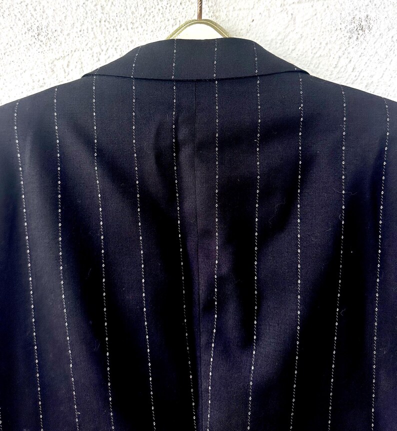 Peut inclure: Un blazer bleu marine fonc&eacute; pr&eacute;sentant un motif &agrave; rayures verticales compos&eacute; de petites lignes pointill&eacute;es claires. Le v&ecirc;tement est pr&eacute;sent&eacute; de dos, montrant un col structur&eacute; et des coutures aux &eacute;paules, suspendu &agrave; un cintre m&eacute;tallique.