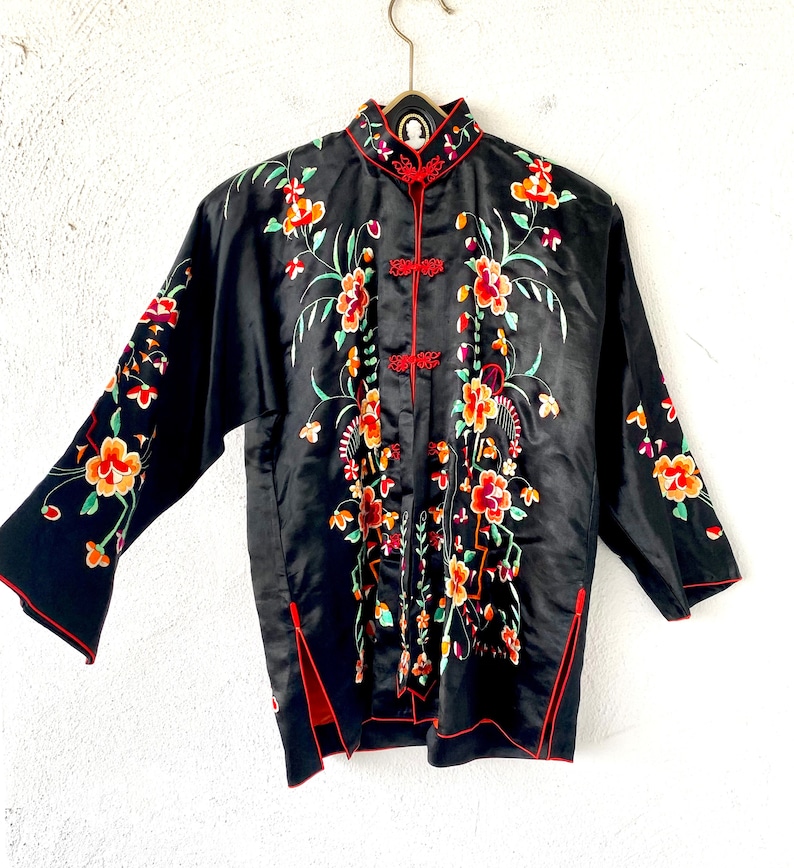 Puede incluir: Chaqueta de seda negra con ribetes rojos y bordados florales. La chaqueta tiene un cuello mandar&iacute;n y cierre de bot&oacute;n.