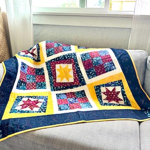 May include: A quilt with a navy blue background and yellow, red, and white stars. The quilt is made of patchwork squares and is folded on a gray couch.