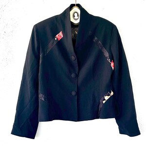 May include: A black blazer with a distinctive design. It has a mandarin collar and three buttons. Floral accents in red, white, and black decorate the front and sleeves. The jacket is suspended from a hanger.