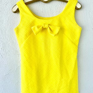 Pu&ograve; includere: Un abito giallo senza maniche con un fiocco sul collo. L'abito ha un tessuto strutturato.