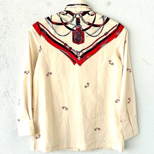 May include: A cream-colored long-sleeved shirt with a red, black, and blue patterned design featuring chains and a keyhole. The pattern is concentrated at the collar and shoulders. The shirt has a button-up front and a small, repeating pattern of red horseshoe shapes throughout.