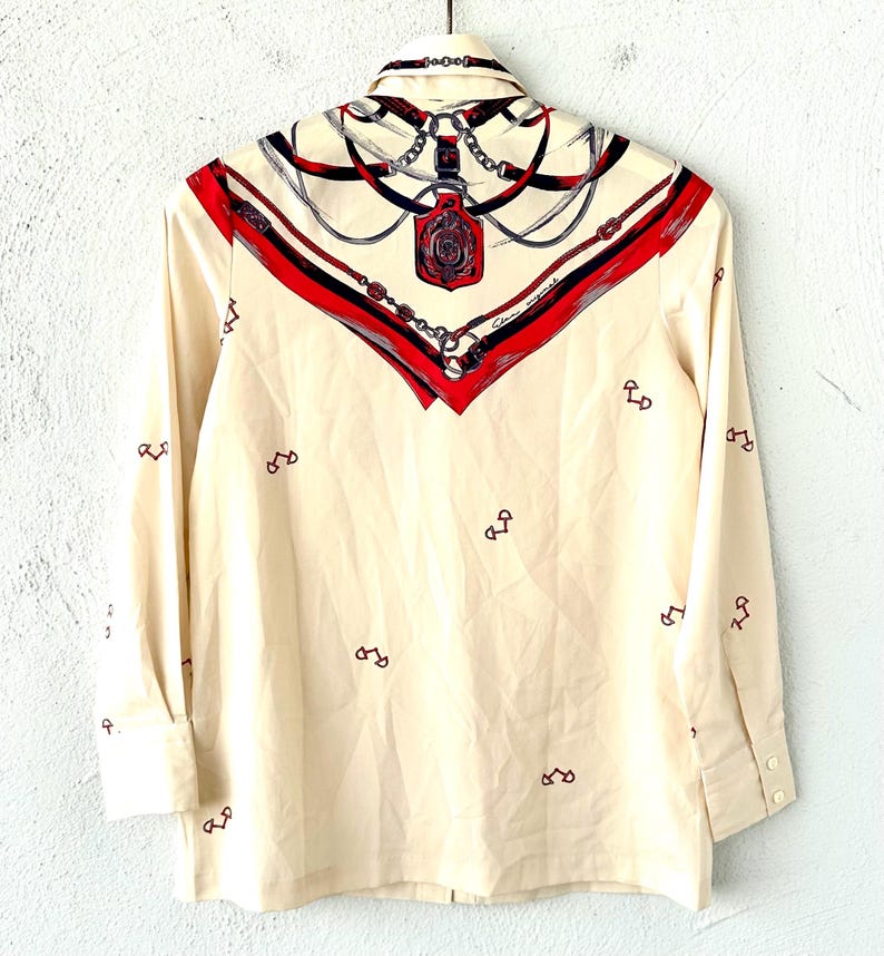 May include: A cream-colored long-sleeved shirt with a red, black, and blue patterned design featuring chains and a keyhole. The pattern is concentrated at the collar and shoulders. The shirt has a button-up front and a small, repeating pattern of red horseshoe shapes throughout.