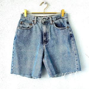 Puede incluir: Pantalones cortos vaqueros azul claro con dobladillo sin rematar, colgados de una percha dorada. Los pantalones cortos tienen el estilo clásico de cinco bolsillos y cierre de botón y cremallera. El denim tiene un aspecto descolorido y desgastado.