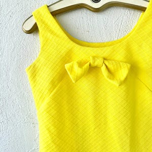 Pu&ograve; includere: Un abito giallo senza maniche con un fiocco sul collo. L'abito ha un tessuto strutturato con un motivo a rombi.