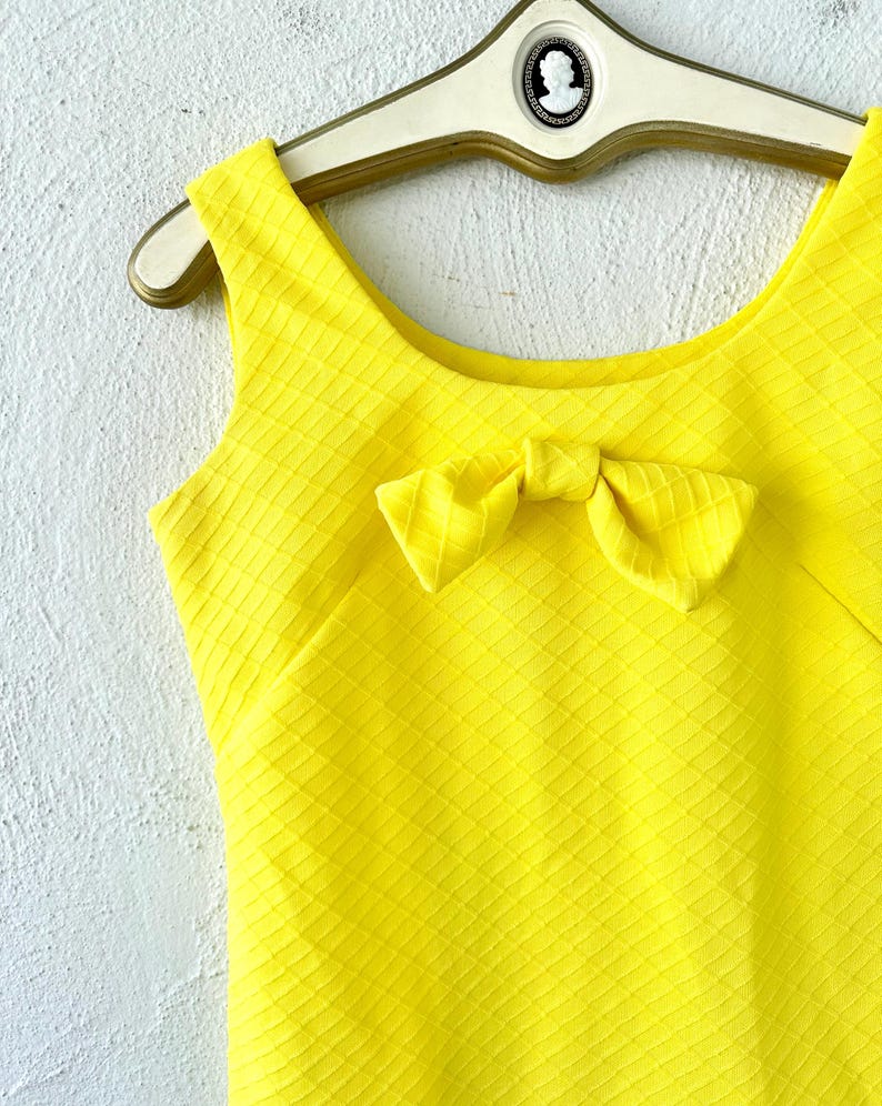 Pu&ograve; includere: Un abito giallo senza maniche con un fiocco sul collo. L'abito ha un tessuto strutturato con un motivo a rombi.