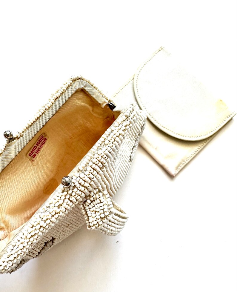 May include: A white beaded clutch purse with a matching white fabric pouch. The clutch has a metal clasp and a label that reads "Hand Made in Belgium".