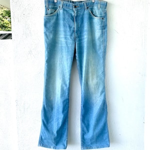 May include: Light blue denim jeans with a classic five-pocket design. The jeans are hanging on a metal hanger against a white wall. The jeans have a faded wash and a slight flare at the bottom.
