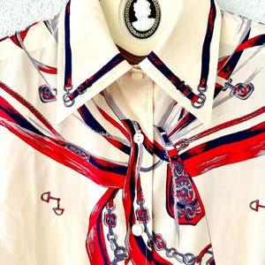 May include: A cream-colored button-down shirt with a red, blue, and gray print of a horse's bridle and reins. The shirt has a pointed collar and a button closure.