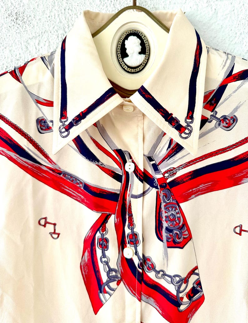 May include: A cream-colored button-down shirt with a red, blue, and gray print of a horse's bridle and reins. The shirt has a pointed collar and a button closure.