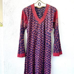 Puede incluir: Vestido de manga larga con cuello en V y mangas acampanadas. El vestido presenta un estampado floral en tonos morados y rojos. El escote y los puños están adornados con una tela roja estampada. El vestido está colgado en una percha vintage.