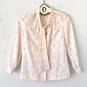 May include: A cream-colored blouse with a star pattern in shades of peach, gray, and black. The long-sleeved top features a tie-neck design and is hanging on a vintage hanger.