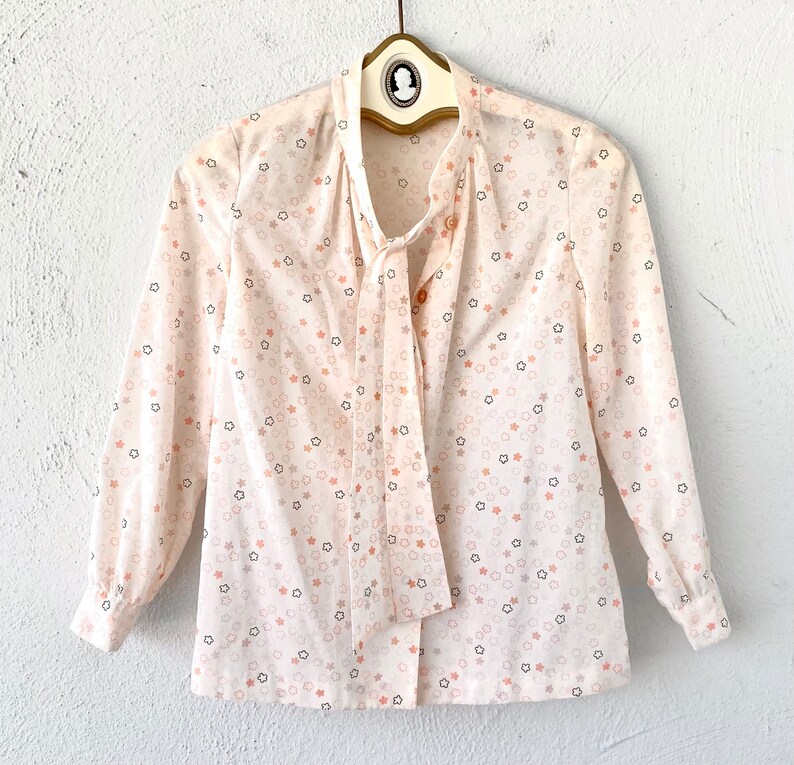 May include: A cream-colored blouse with a star pattern in shades of peach, gray, and black. The long-sleeved top features a tie-neck design and is hanging on a vintage hanger.
