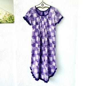 May include: A knee-length purple and white dress featuring a patchwork-style floral and leaf print. The garment has short sleeves with ruffled edges, a scoop neckline, a central vertical pleated panel with three buttons, and a ruffled hemline.