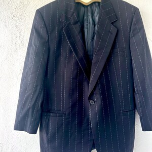 Peut inclure: Un blazer bleu marine fonc&eacute; orn&eacute; d'un motif &agrave; fines rayures blanches avec de petits points. La veste pr&eacute;sente un revers crant&eacute;, une fermeture &agrave; un bouton, deux poches lat&eacute;rales &agrave; rabat et des manches longues, suspendue &agrave; un cintre avec un cam&eacute;e.