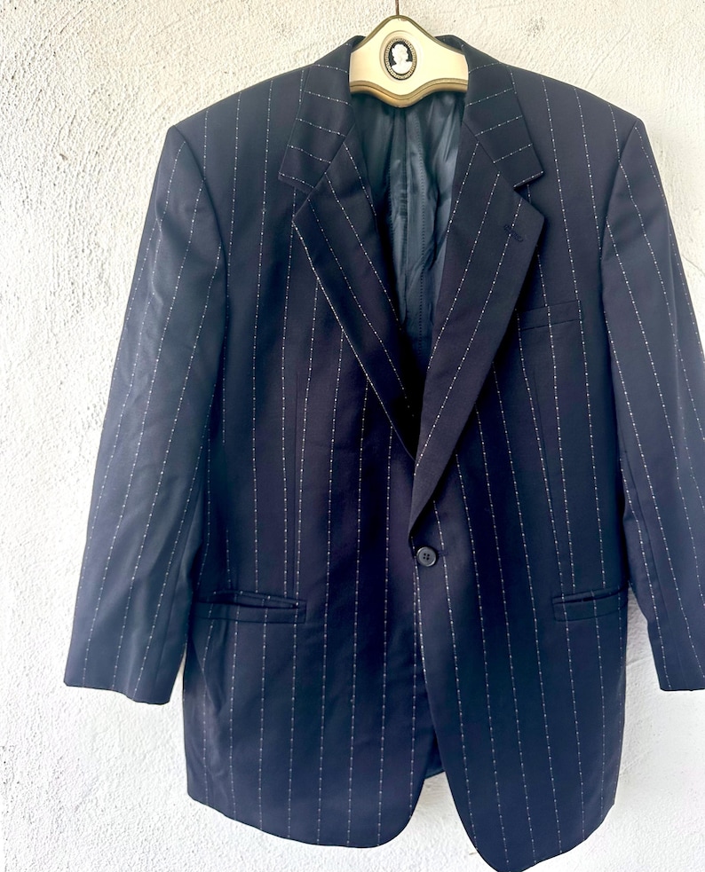 Peut inclure: Un blazer bleu marine fonc&eacute; orn&eacute; d'un motif &agrave; fines rayures blanches avec de petits points. La veste pr&eacute;sente un revers crant&eacute;, une fermeture &agrave; un bouton, deux poches lat&eacute;rales &agrave; rabat et des manches longues, suspendue &agrave; un cintre avec un cam&eacute;e.