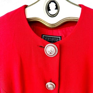 Puede incluir: Una chaqueta roja brillante con dos botones plateados decorativos con un dise&ntilde;o de cabeza de le&oacute;n. La chaqueta est&aacute; colgada en una percha blanca con un dise&ntilde;o de camafeo en blanco y negro. La etiqueta dice "VERSUS".