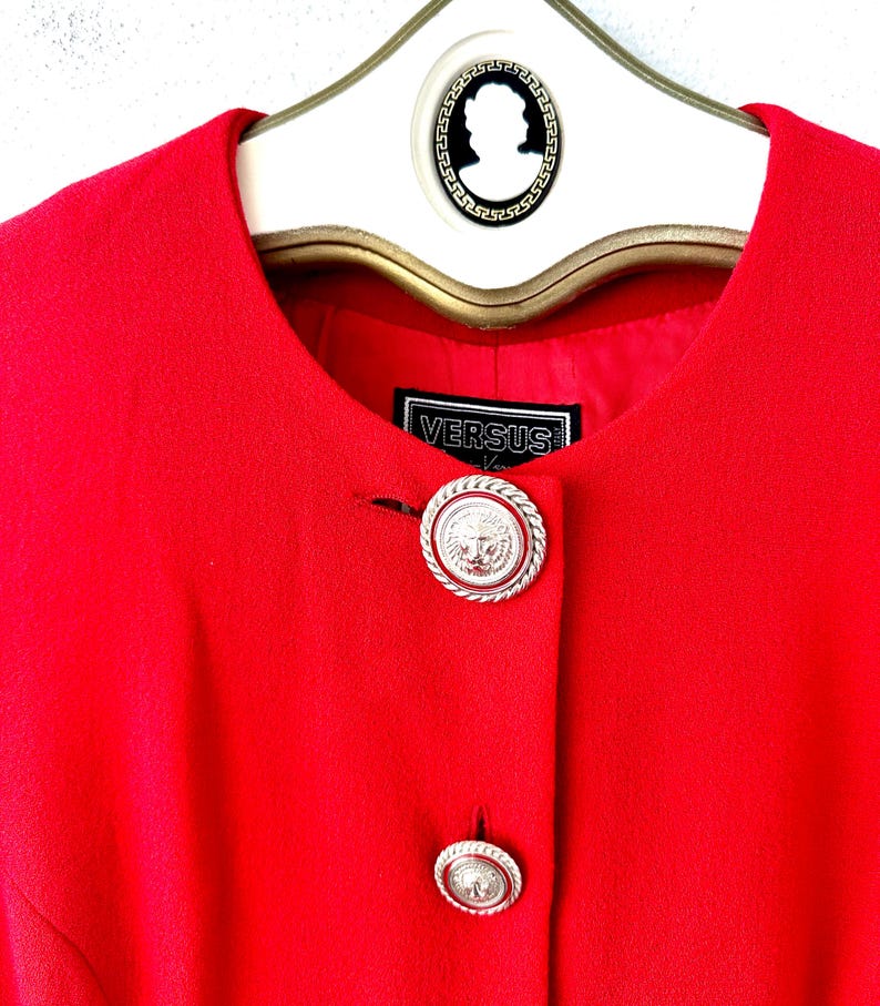Puede incluir: Una chaqueta roja brillante con dos botones plateados decorativos con un dise&ntilde;o de cabeza de le&oacute;n. La chaqueta est&aacute; colgada en una percha blanca con un dise&ntilde;o de camafeo en blanco y negro. La etiqueta dice "VERSUS".
