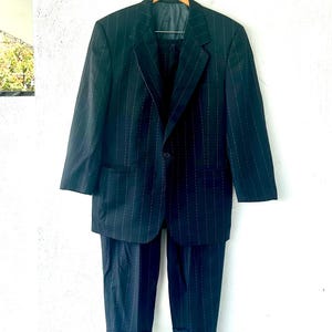 Peut inclure: Un ensemble de costume bleu marine fonc&eacute; avec un motif &agrave; fines rayures blanches. Il se compose d'une veste &agrave; simple boutonnage avec des revers crant&eacute;s et un bouton unique, accompagn&eacute;e d'un pantalon assorti avec un ourlet &agrave; revers.