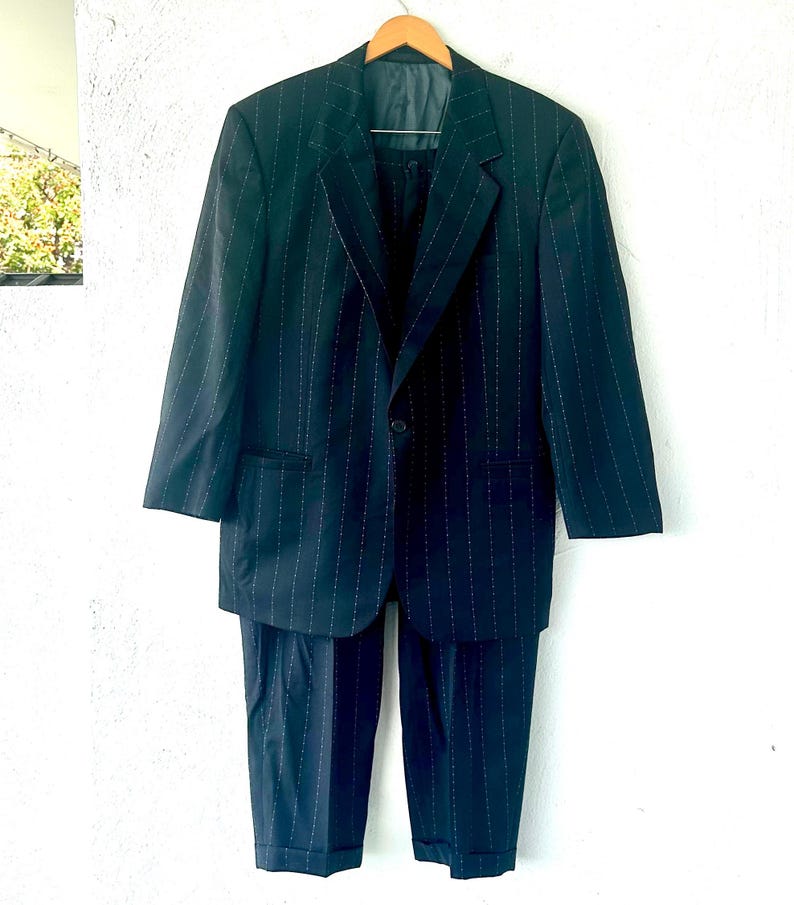 Peut inclure: Un ensemble de costume bleu marine fonc&eacute; avec un motif &agrave; fines rayures blanches. Il se compose d'une veste &agrave; simple boutonnage avec des revers crant&eacute;s et un bouton unique, accompagn&eacute;e d'un pantalon assorti avec un ourlet &agrave; revers.
