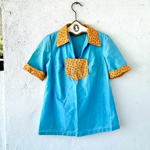 Puede incluir: Una camisa de manga corta azul claro con un cuello y un bolsillo con estampado amarillo y rojo.