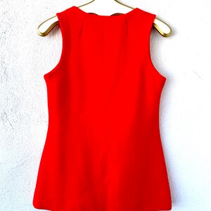 May include: A sleeveless, bright red top hangs on a gold hanger against a white wall. The top has a textured fabric and a slightly flared shape. The neckline has a subtle, scalloped design.