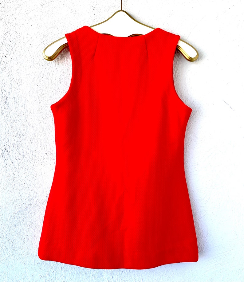 May include: A sleeveless, bright red top hangs on a gold hanger against a white wall. The top has a textured fabric and a slightly flared shape. The neckline has a subtle, scalloped design.