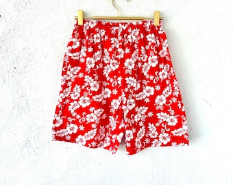 May include: Red shorts with a white floral pattern, hanging from a gold-colored hanger. The shorts feature an elastic waistband and a vibrant all-over print of white hibiscus flowers and smaller star-shaped blossoms.