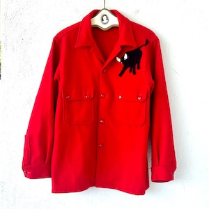 May include: A bright red button-down shirt with two chest pockets and a black bull applique on the left chest. The long-sleeved shirt is hanging on a hanger against a white wall.