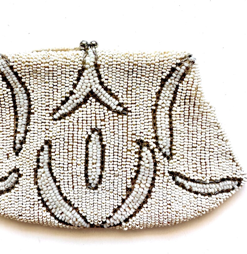 May include: A white beaded clutch purse with a silver clasp. The purse has a geometric design with a repeating pattern of curved lines.