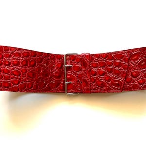 Op de afbeelding: Een brede, rode riem met een krokodillenleertextuur. De riem heeft een zilverkleurige, rechthoekige gesp. De riem is ontworpen om de taille te accentueren en een gedurfd accent aan elke outfit toe te voegen.