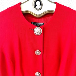 Puede incluir: Una blazer rojo brillante con cuello redondo y tres botones plateados. Los botones tienen un dise&ntilde;o detallado. La blazer tiene la palabra "VERSUS" en una peque&ntilde;a etiqueta. La blazer est&aacute; colgada en una percha blanca con una parte superior decorativa.