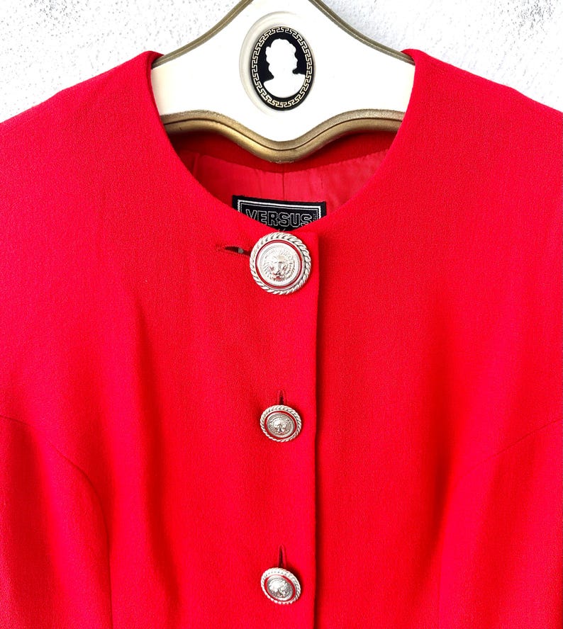 Puede incluir: Una blazer rojo brillante con cuello redondo y tres botones plateados. Los botones tienen un dise&ntilde;o detallado. La blazer tiene la palabra "VERSUS" en una peque&ntilde;a etiqueta. La blazer est&aacute; colgada en una percha blanca con una parte superior decorativa.
