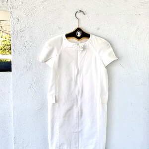 May include: A white short-sleeved dress with a round neckline and a button closure at the neck. The dress has a fitted silhouette and a straight hemline.