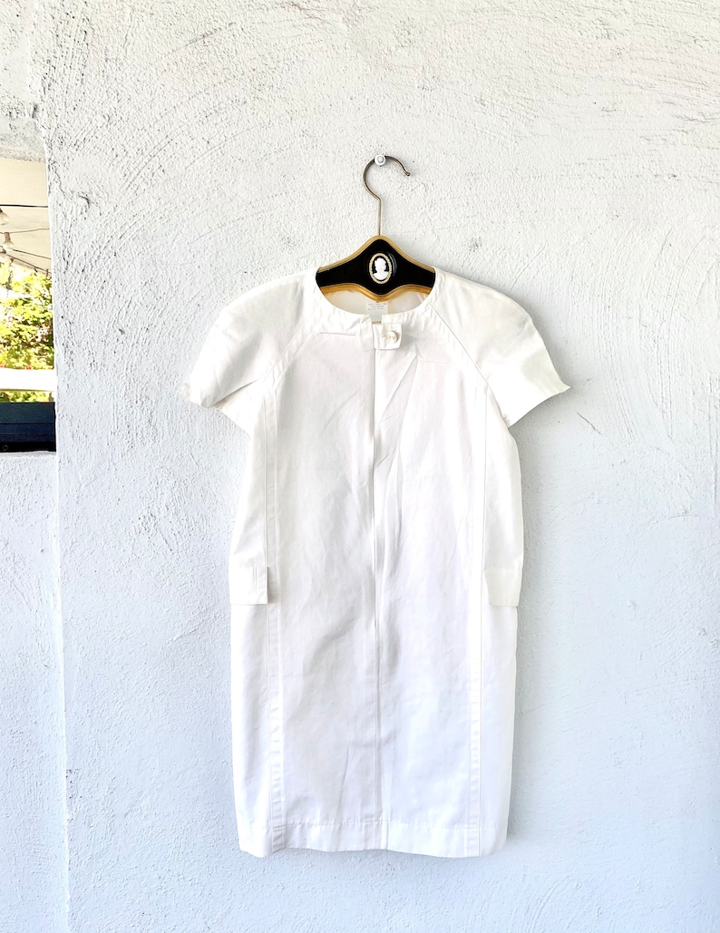 May include: A white short-sleeved dress with a round neckline and a button closure at the neck. The dress has a fitted silhouette and a straight hemline.