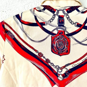 May include: A white silk shirt with a red, blue, and gray pattern of chains, belts, and a decorative buckle. The shirt has the text "Alan origina" printed on the bottom right corner.
