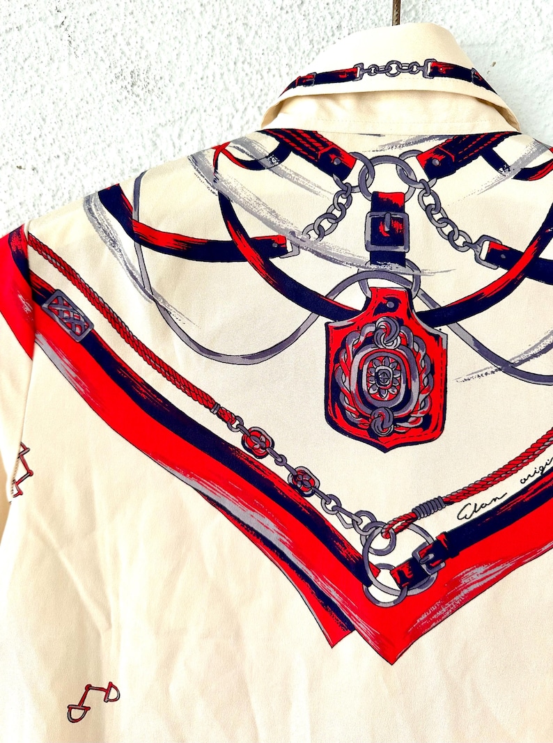 May include: A white silk shirt with a red, blue, and gray pattern of chains, belts, and a decorative buckle. The shirt has the text "Alan origina" printed on the bottom right corner.