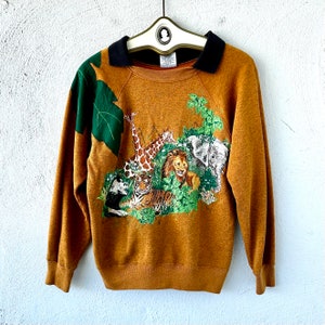 May include: A brown sweatshirt with a black collar and a colorful jungle animal print. The print features a giraffe, a tiger, a lion, and an elephant.
