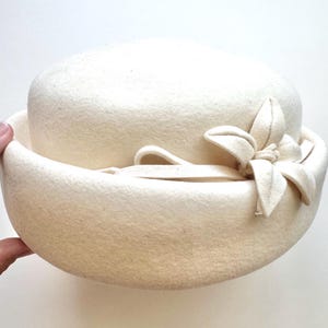 May include: Cream-colored cloche hat with a rounded crown and a wide brim. A fabric flower with leaf-like details adorns the side. The hat is made of a soft, felt-like material, offering a vintage aesthetic. A classic accessory.