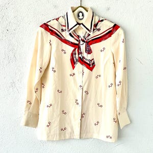 May include: A cream-colored long-sleeved button-down shirt with a red, blue, and white patterned scarf collar. The shirt is covered in a repeating pattern of small red horseshoes.