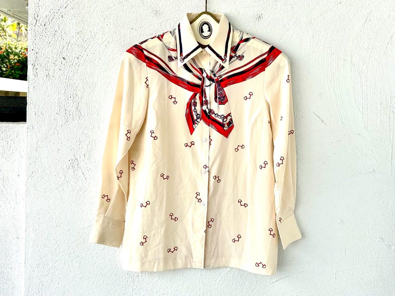 May include: A cream-colored long-sleeved button-down shirt with a red, blue, and white patterned scarf collar. The shirt is covered in a repeating pattern of small red horseshoes.