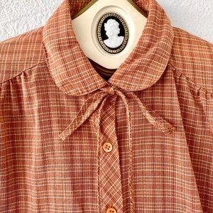 May include: A brown and white plaid shirt with a peter pan collar and a bow tie. The shirt has a button-up front and long sleeves.
