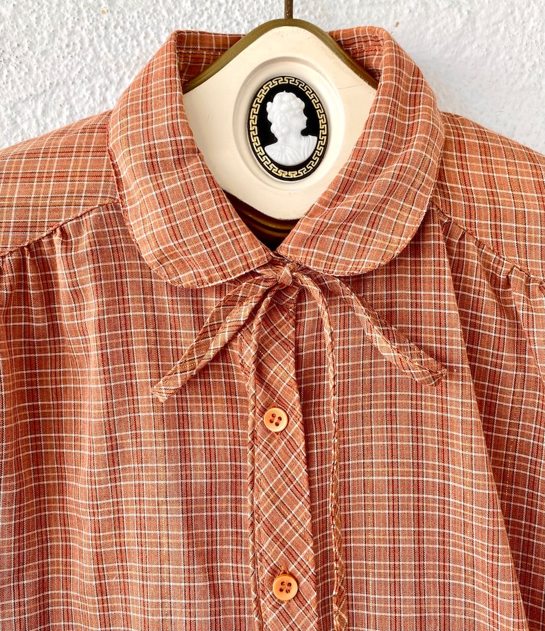 May include: A brown and white plaid shirt with a peter pan collar and a bow tie. The shirt has a button-up front and long sleeves.